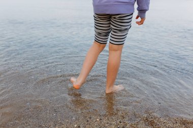 close up on barefeet child legs in striped pants standing on stone beach at the lake