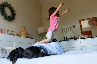 Mavi pijamalı iki kız yatağa uzanıyor ve diğer kız kardeş üzerinden atlıyor.