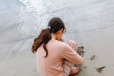 back view of sitting girl child with long hair drawing on the sand beach