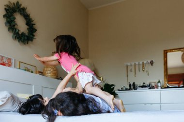 Mavi pijamalı iki kız yatağa uzanıyor ve diğer kız kardeş üzerinden atlıyor.