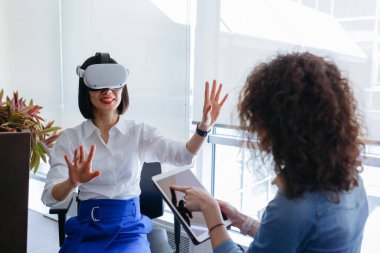 Kısa siyah saçlı kadın sanal gerçeklik görüntüleyicisi takıyor ve kıvırcık saçlı meslektaşı onu tabletle takip ediyor.