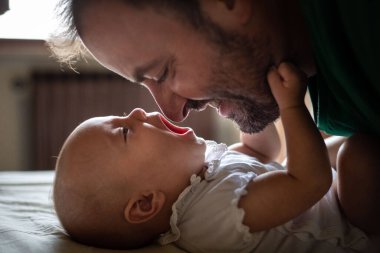 Baba yüzü sakallı, beyaz bluzlu bebeğini yatakta yatarken seyrediyor.