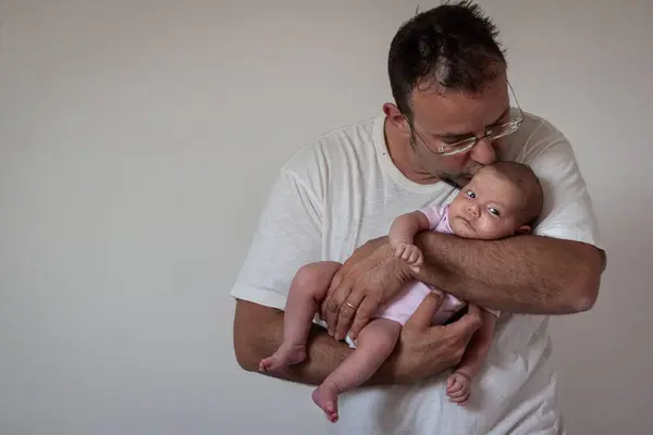 father in white t-shirt holding newborn baby in his arms and kissing the daughter in pink