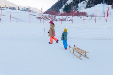 İki çocuk parlak kış elbiseleriyle kar yamacında ahşap kızağı çekiyor.