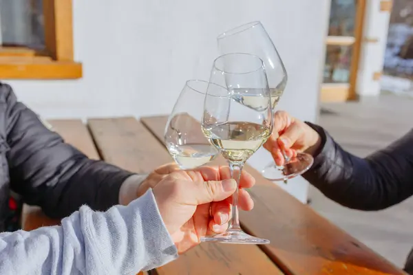 close up on hands of three people in warm jackets holding glasses of sparkling wine prosecco sitting at wooden table outside white house