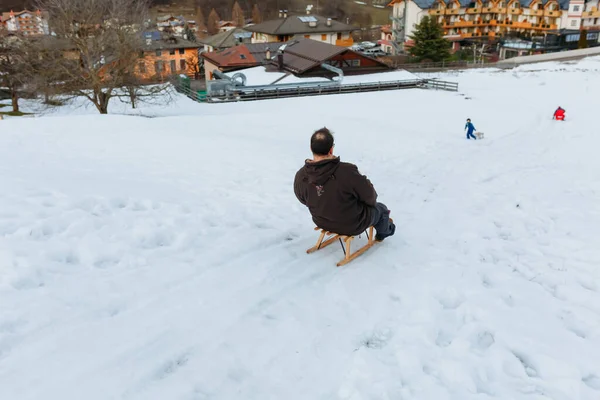 rear view of mid aged man in brown jacket sitting on wooden sled going down the snow hill