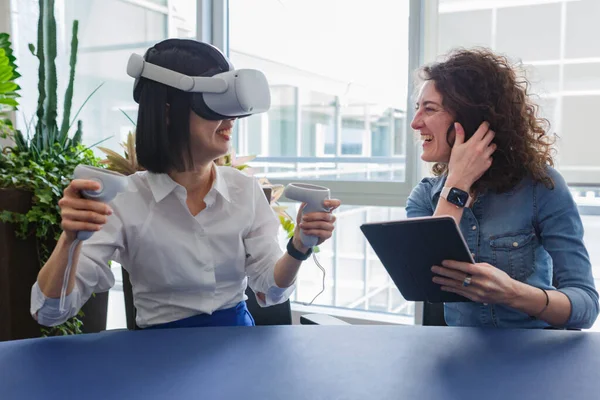 upper view of two happy women sitting at the table in the office and working with virtual reality viewer and tablet