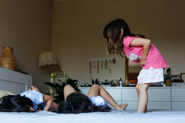 Mavi pijamalı iki kız yatağa uzanıyor ve diğer kız kardeş atlamaya hazırlanıyor.