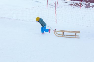 Küçük kız parlak kış kıyafetleriyle kar yamacında ahşap kızağı çekiyor