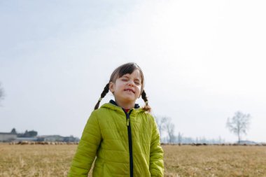 Yeşil ceketli, saçları örgülü, tarlada gözleri kapalı, gülümseyen küçük bir kız.