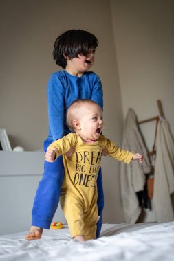 İkisi yatakta mavi ve sarı elbiseli çocuklar mutlu bir şekilde izliyorlar.