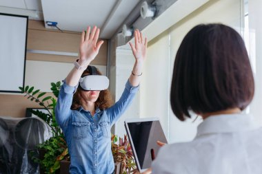 Kıvırcık kahverengi saçlı ve kot gömlekli bir kadın ofiste VR deniyor. Önde Asyalı meslektaşı tabletle çalışıyor.