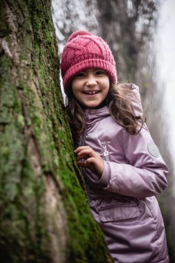 Ağacın arkasına saklanan pembe şapkalı gülümseyen kızın portresi.