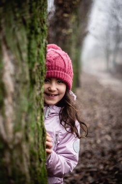 Ağacın arkasına saklanan pembe şapkalı gülümseyen kızın portresi.