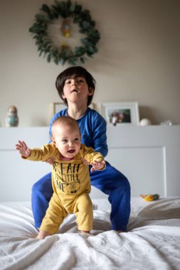 İkisi yatakta duruyor. Mavi ve sarı elbiseli çocuklar ellerinde küçük bir bebekle oynuyorlar.