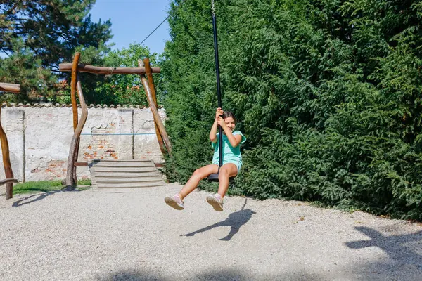 smiling girl in green outfit sitting on a pulley on a sunny day in the park