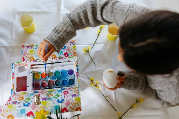 upper view of child in grey sweater sitting at the white table and coloring the egg with yellow paint