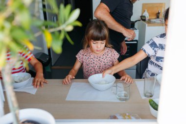 Üç kardeş evde yemek yerken, babam arka planda yemek pişiriyor.