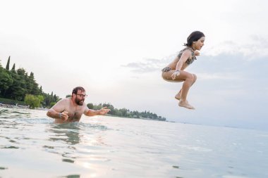 Gün batımında babanın omuzlarından suya atlayan mayo giymiş kızın yan görüntüsü.