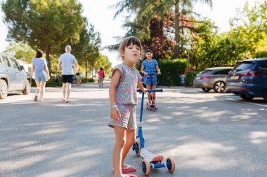 Güneşli bir günde kız kardeşiyle mobiletin önünde duran renkli elbiseli çocuğun düşük açılı görüntüsü.