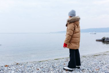 Kışlık ceketli ve gri şapkalı bir çocuğun çakıl taşlı kumsalda dikiliş görüntüsü