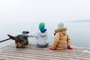 Sisli bir günde bir iskelede köpekle oturan iki kızın arka görüntüsü.