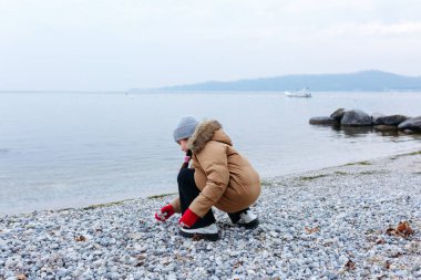 Kışlık ceketli, gri şapkalı, taşlarla oynayan çakıl taşlı plaj çocuğunun arka manzarası.