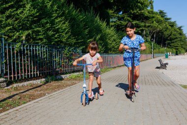 İki çocuk parkta güneşli bir günde scooter kullanıyor.
