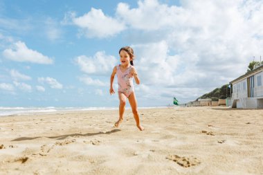 Mavi gökyüzüne karşı kumlu sahilde koşan pembe mayo giymiş gülümseyen küçük kızın alçak açılı görüntüsü.