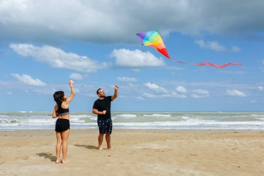Baba ve kızı kumlu bir sahilde gökkuşağı uçurtması uçuruyorlar güneşli bir günde denizin kenarında duruyorlar.