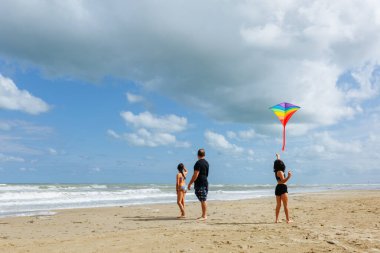 Babamın iki kızıyla güneşli bir günde kumsalda gökkuşağı uçurtması uçururken, denizin kenarında dururken geniş açılı görüntüsü.