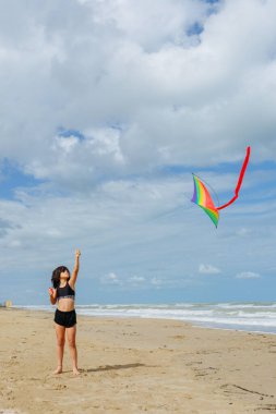 Geniş açı, kumsalda çok renkli bir uçurtma uçuran koyu saçlı bir kız görüntüsü.