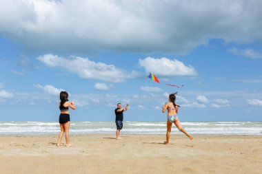Güneşli bir günde kumsalda gökkuşağı uçurtması uçuran iki kızı olan babayı deniz kenarında oynarken geniş açılı görmek.