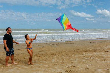 Baba ve kızı kumlu bir sahilde gökkuşağı uçurtması uçuruyorlar güneşli bir günde denizin önünde duruyorlar