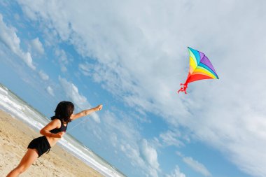 Sahilde çok renkli bir uçurtma uçuran koyu renk saçlı kızın düşük açılı görüntüsü.