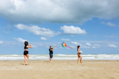 Güneşli bir günde kumsalda gökkuşağı uçurtması uçuran iki kızı olan bir babanın deniz kenarında oynadığı geniş açılı bir manzara.