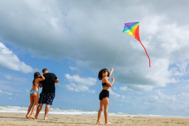 Güneşli bir günde gökkuşağı uçurtması uçuran iki kızı olan bir babanın arka görüntüsü.