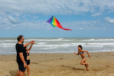 Baba ve kızı, güneşli bir günde kumsalda gökkuşağı uçurtması uçuruyorlardı. Denizin önünde duruyorlardı. Bir kız daha yanlarında koşuyordu.