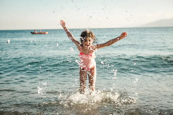 Çizgili mayo giymiş, deniz suyuyla oynayan küçük bir kız.