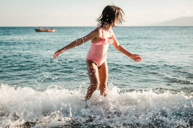 Çizgili mayolu küçük kız denizde dalgalarla oynuyor.