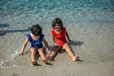Parlak mayo giymiş iki kız ve yüzlerinde güneş kremi suyun yanında oturmuş gülümsüyor.
