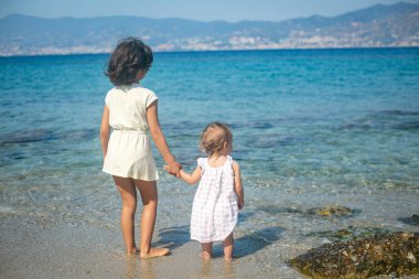 Küçük kardeşin el ele tutuşup sahilde bir arada duran ablasının arkadan görünüşü. Öndeki denize bakıyor.