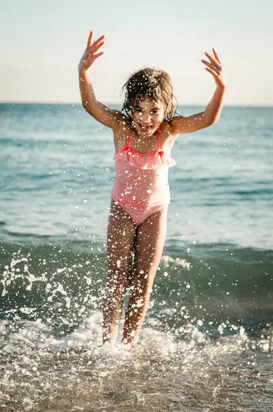 Çizgili mayo giymiş, denizde oynayan, su sıçratan küçük bir kız resmi.