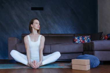 Yoga pozisyonunda oturan kadın evinde kanepenin yanında egzersiz yapıyor.