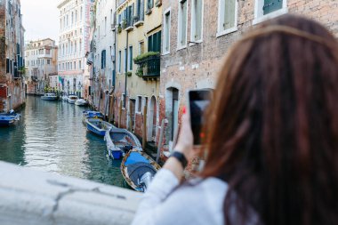 Koyu renk saçlı, manikürlü, Venedik Kanalı 'nın cep telefonuyla fotoğrafını çeken bir kadının arka görüntüsü.