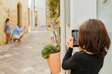 Güneşli bir günde dar bir sokakta kız kardeşinin fotoğrafını çeken bir kızın cep telefonu görüntüsü.