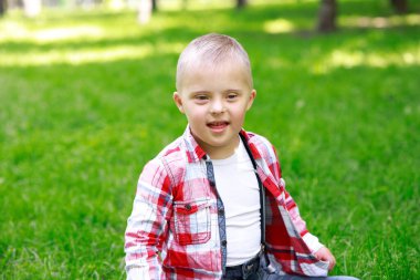 Parkta Down Sendromlu bir çocuk var. Yeşilliğin arka planına karşı. Çocuk gülümsüyor.