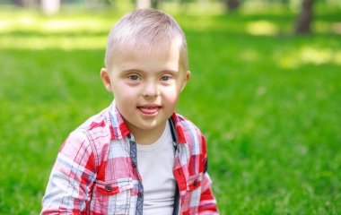 Parkta Down Sendromlu bir çocuk var. Yeşilliğin arka planına karşı. Çocuk gülümsüyor.