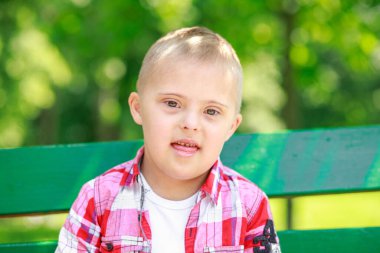 Parkta Down Sendromlu bir çocuk var. Yeşilliğin arka planına karşı. Çocuk gülümsüyor.