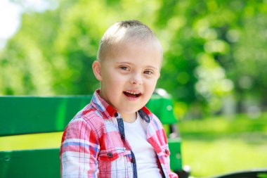 Parkta Down Sendromlu bir çocuk var. Yeşilliğin arka planına karşı. Çocuk gülümsüyor.